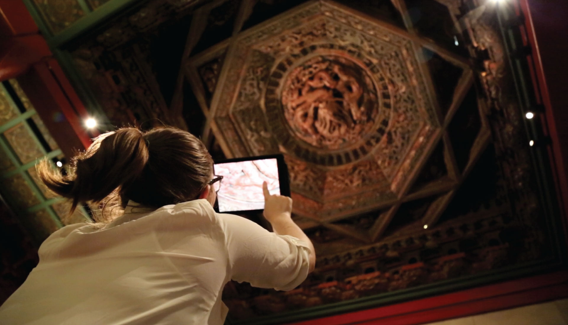 A woman standing under the temple ceiling holding the iPad above her head to explore the ceiling. 